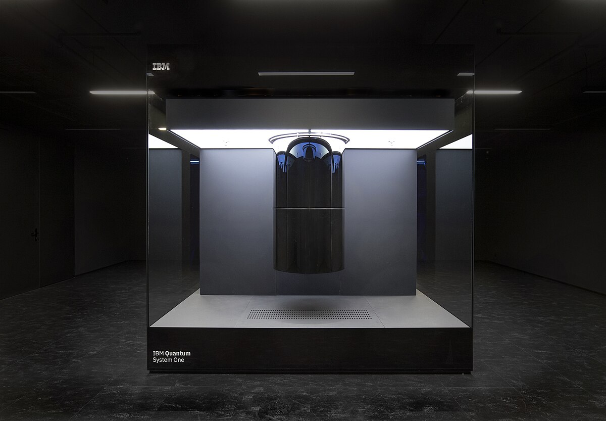 The IBM Q System One, the first circuit-based commercial quantum computer, is displayed within a large glass enclosure. The system, developed in 2019, features a cylindrical quantum processor unit suspended in the center of the case.