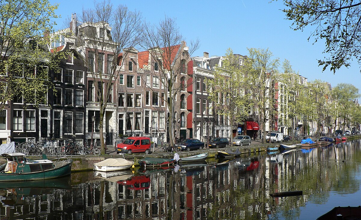A canal in Amsterdam with traditional Dutch houses lining its banks and boats moored along the water's edge. The buildings feature gabled roofs and narrow facades, characteristic of Amsterdam's architecture.