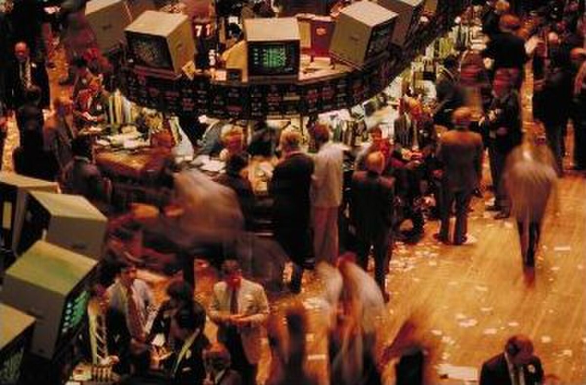 Traders are actively working on the floor of a stock exchange, with some figures blurred due to motion, indicating a busy environment. The scene features multiple trading desks with computer monitors and a large, circular overhead display.