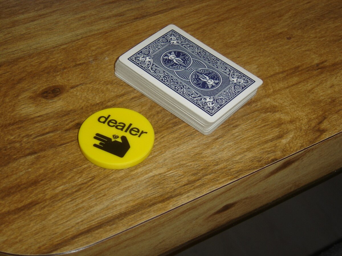 Yellow poker dealer button chip and a deck of blue playing cards resting on a wooden table surface. The circular chip features the word "dealer" in black lowercase text above a graphic icon of a hand holding a playing card.