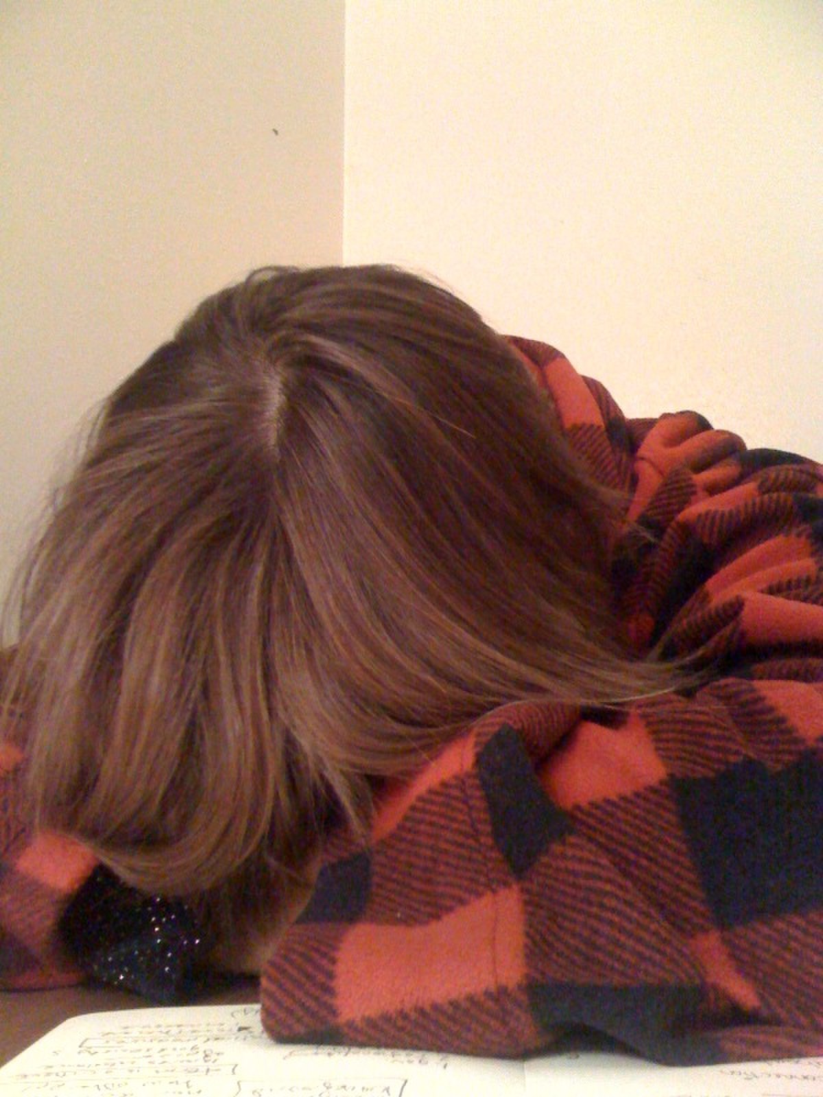 A student is shown with their head buried in their arms on a desk, covered by a red and black plaid blanket, indicating stress or exhaustion from homework. A notebook with handwritten text is visible beneath their arms.