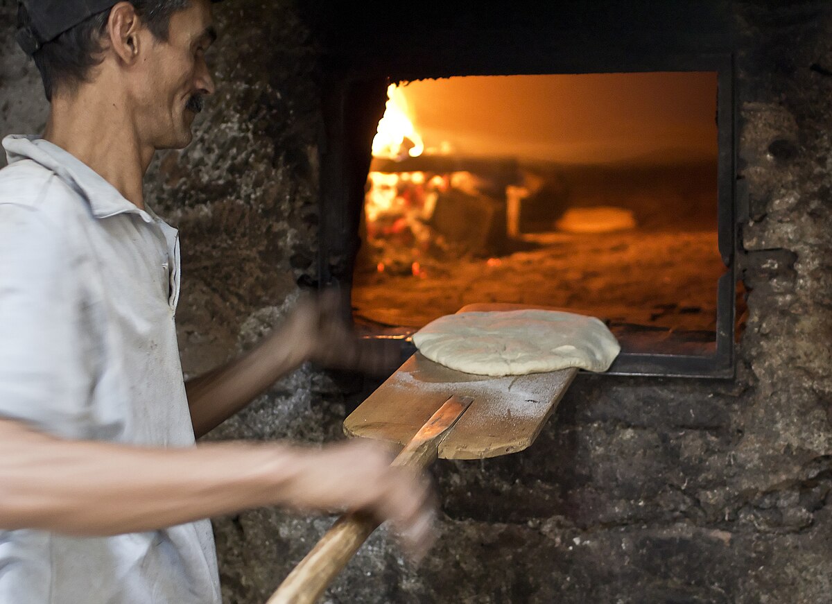 A person baking bread in a traditional communal oven, using a long wooden peel to place a flatbread into the fiery oven. This illustrates secondary food processing.