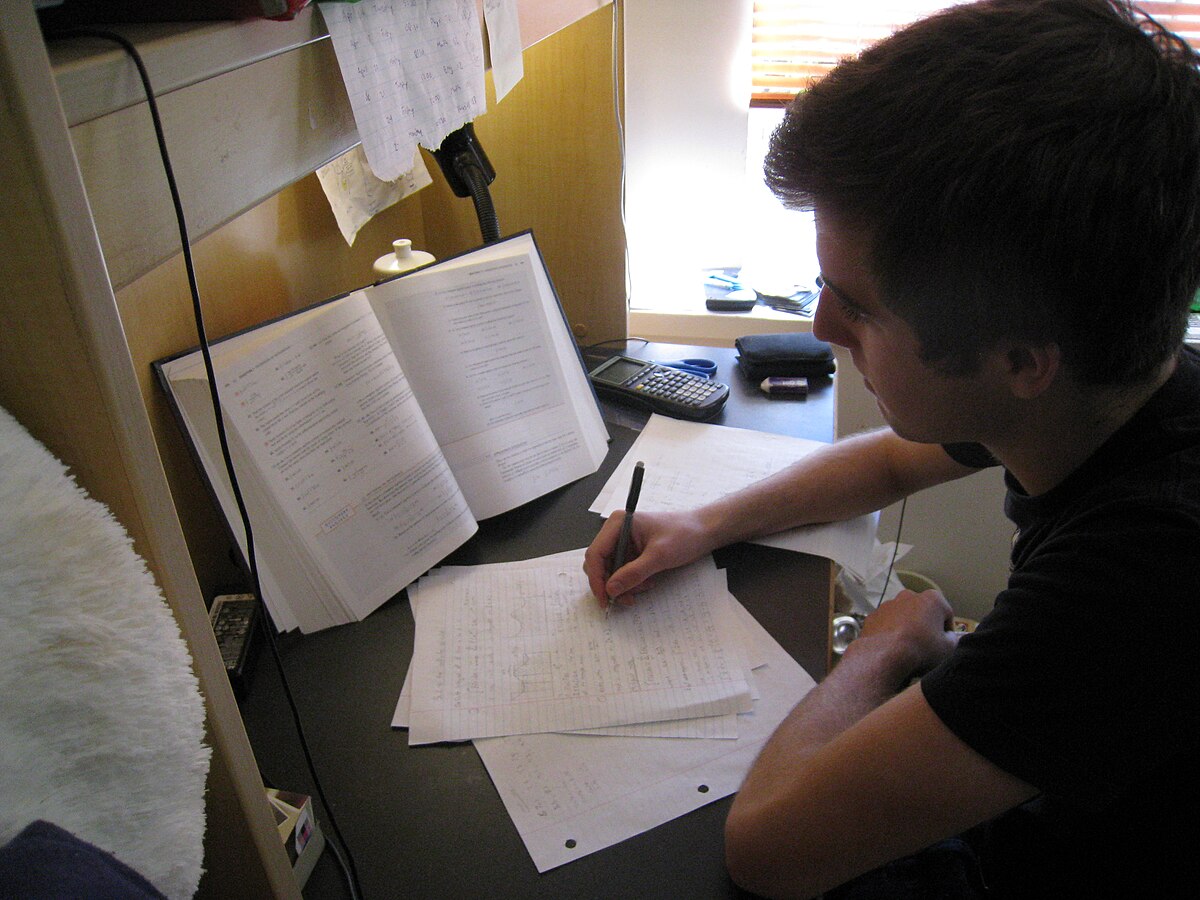 A student studying at a desk, writing on papers with an open textbook and a calculator nearby. The scene illustrates the importance of preparing for tests in similar contexts to aid recall.