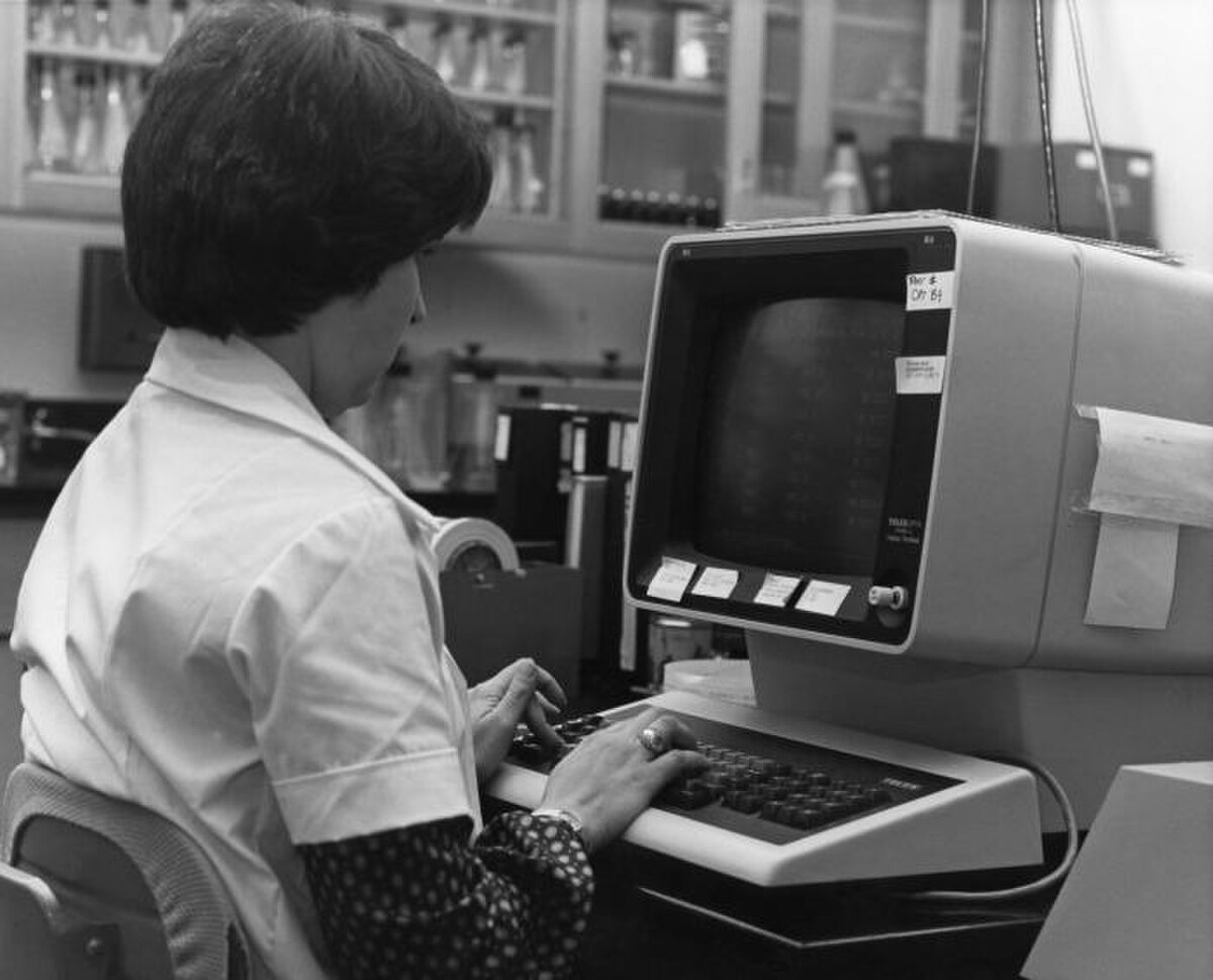 A worker at the Centers for Disease Control in the 1980s is shown typing on a computer keyboard, with a CRT monitor displaying text. The worker is wearing a lab coat, and laboratory equipment is visible in the background.