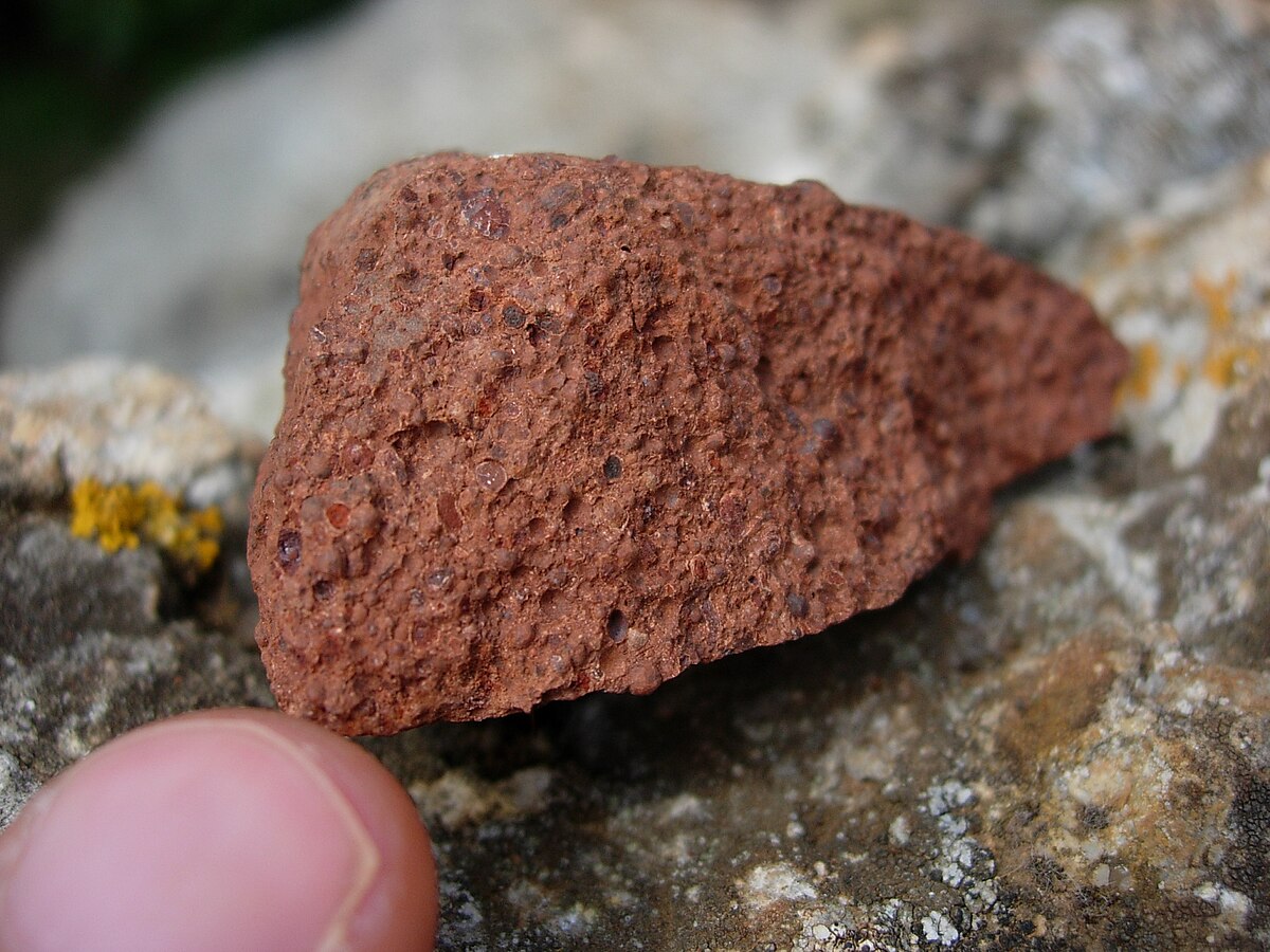 Close-up of a piece of bauxite, a major aluminium ore, held between a thumb and forefinger. The rock is reddish-brown and porous, with the color attributed to the presence of iron oxide minerals.