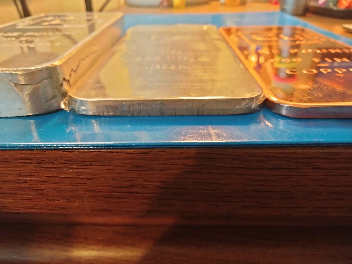 Three 1.0 pound (0.454 kg) bullion ingots of aluminium, iron, and copper are shown side-by-side on a blue tray. The ingots are etched with their scientific details, with the copper ingot on the right clearly showing "ONE POUND FINE COPPER" and the aluminum ingot in the middle showing "ALUMINIUM" and "999 FINE."