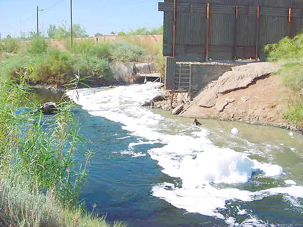 The New River flowing from Mexicali, Baja California, into the Imperial Valley, showing foamy, discolored water entering a larger body of water near a building.