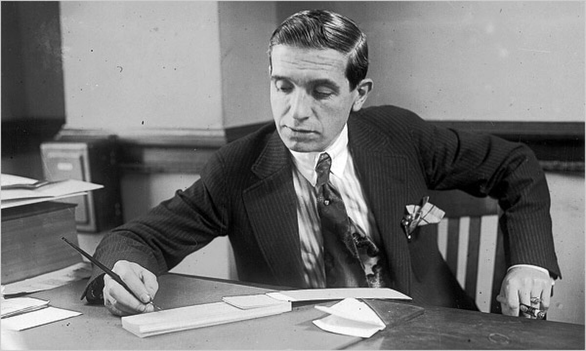 Black and white photograph of Charles Ponzi, taken in 1920. He is seated at a desk, wearing a suit and tie, holding a pen and looking down at papers.