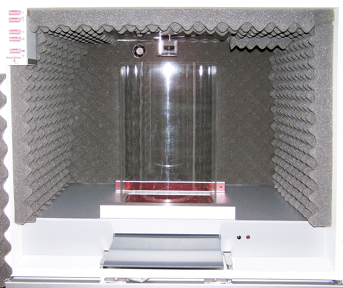 A fear conditioning apparatus for mice is shown, consisting of a sound-attenuating chamber lined with foam, containing a clear cylindrical enclosure on a metal grid floor. The apparatus is equipped with a sound, a foot shock mechanism, and an activity sensor with photobeams to measure freezing behavior.