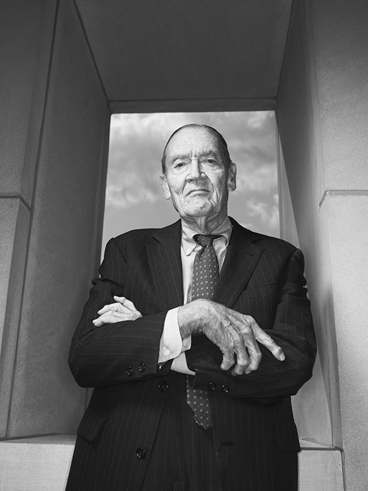 Black and white portrait of John C. Bogle, an older man in a suit and tie, with his arms crossed, standing in front of a window with a cloudy sky visible. The image captures him with a serious expression.