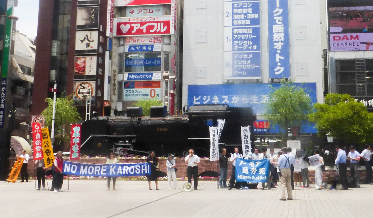 A "No More Karoshi" protest in Tokyo, Japan, on June 13, 2018, with people holding banners and signs in front of a black steam locomotive. Large buildings with Japanese advertisements are visible in the background.