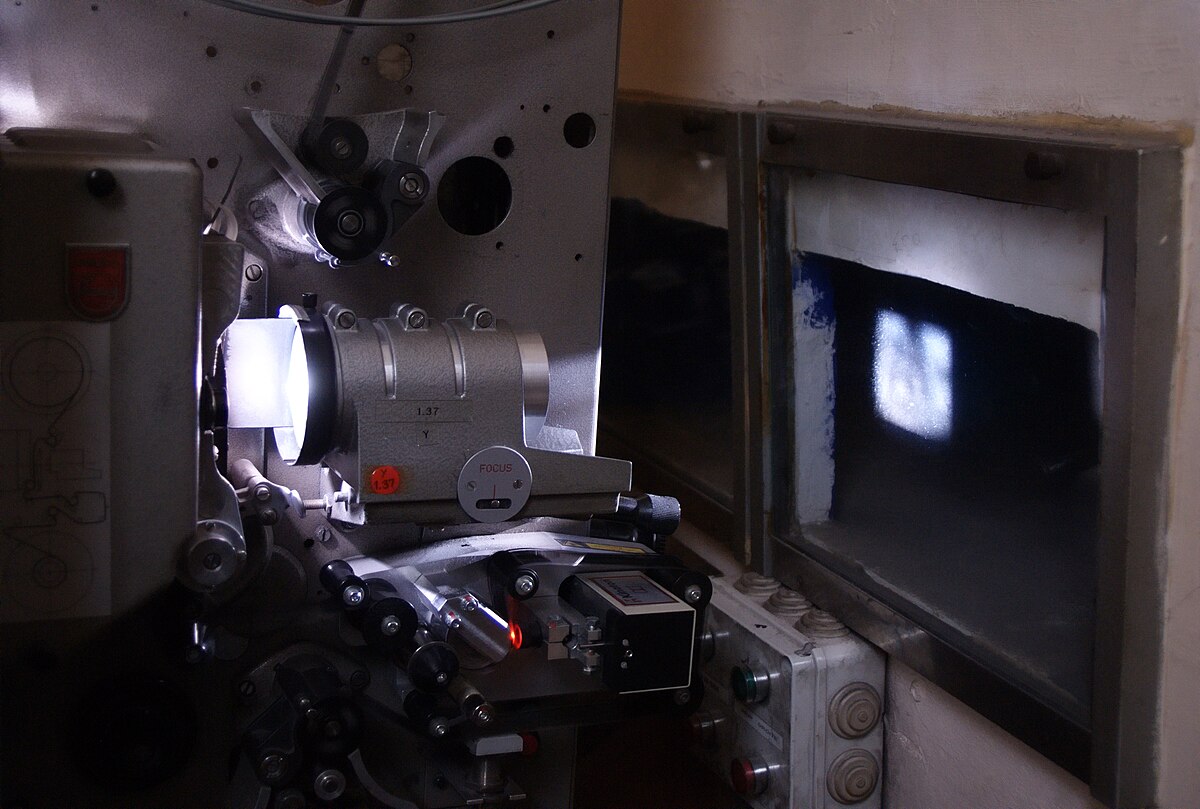 A 35mm movie projector in operation, with a bright light beam emanating from the lens and projecting an image onto a small screen or window. The intricate mechanical components of the projector are visible.