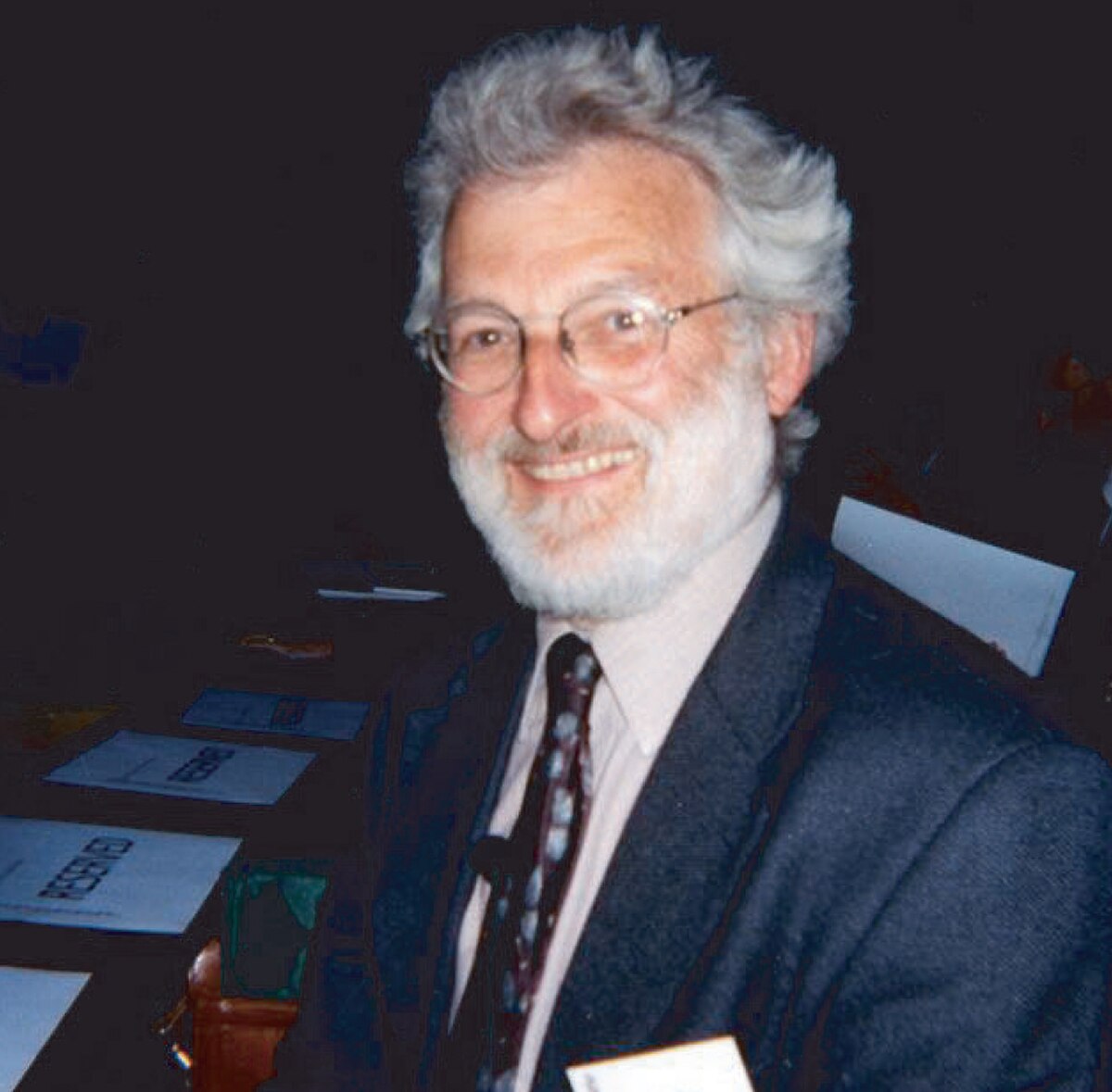 Portrait of John Sulston, a man with gray hair and a beard, wearing glasses, a dark suit jacket, and a patterned tie, smiling at the camera. He was awarded the Nobel Prize in Medicine in 2002 for his research on apoptosis.
