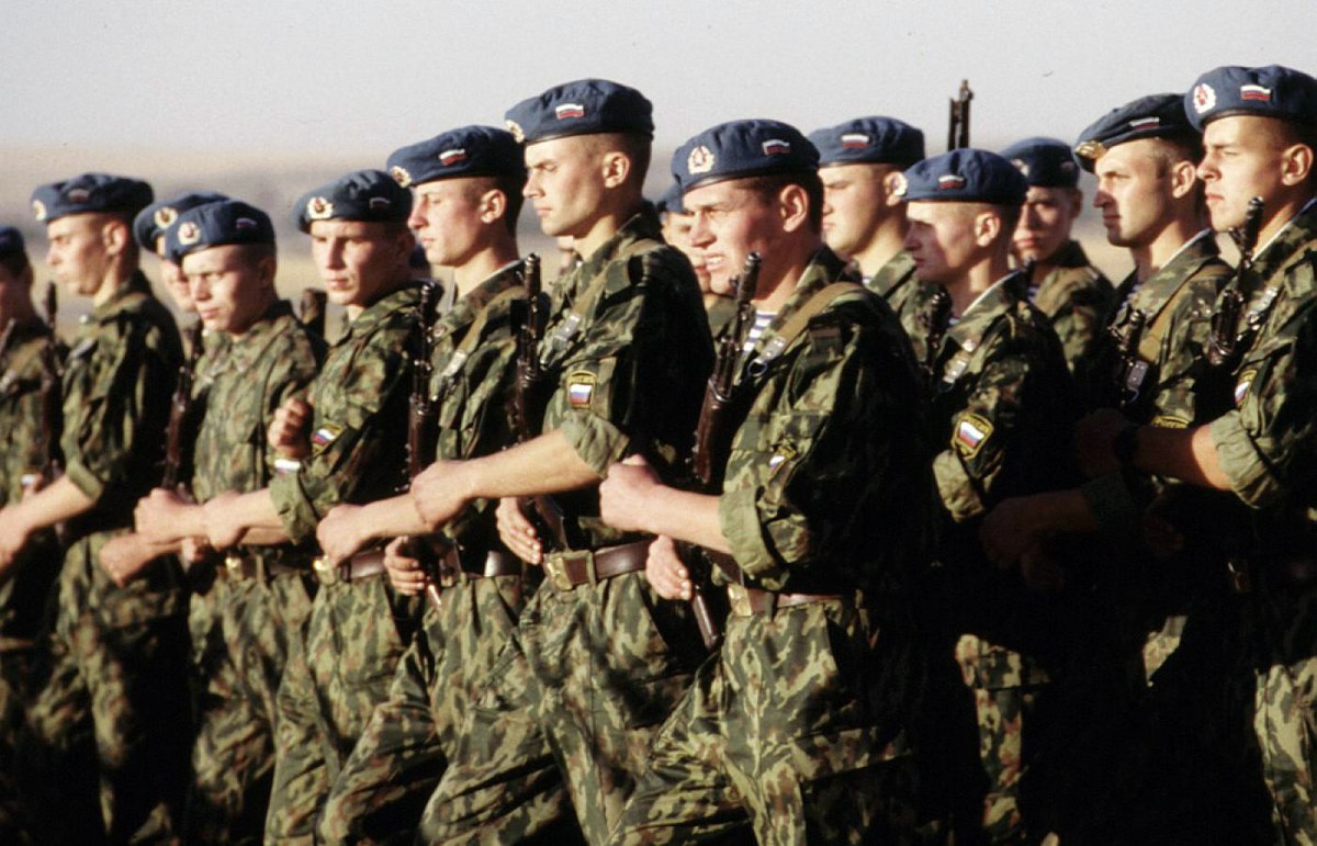 A line of Russian paratroopers in camouflage uniforms and blue berets stands at attention in Kazakhstan. They are equipped with rifles and appear to be participating in a military exercise or parade.