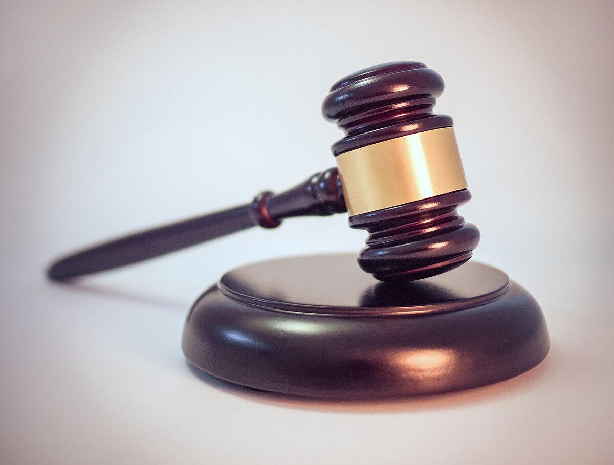 A wooden legal gavel with a brass band on its head, resting on a matching wooden sound block. The gavel and block are dark brown with a polished finish.
