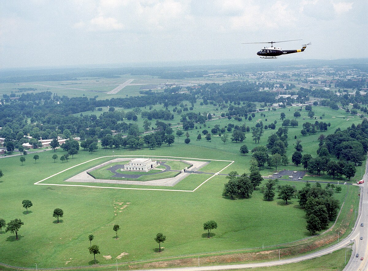 An aerial view of the U.S. Gold Bullion Depository at Fort Knox, Kentucky, with an UH-1 Iroquois helicopter flying overhead. The depository building is a small, fortified structure surrounded by a large, manicured lawn and security fences.