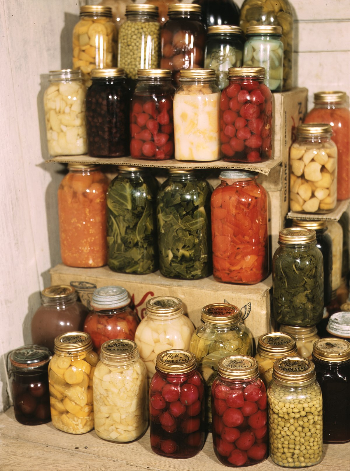 Numerous glass jars filled with various preserved foods, including fruits and vegetables, are stacked on shelves. The jars contain items such as green beans, cherries, corn, and other colorful produce, indicating a home canning or food preservation effort.