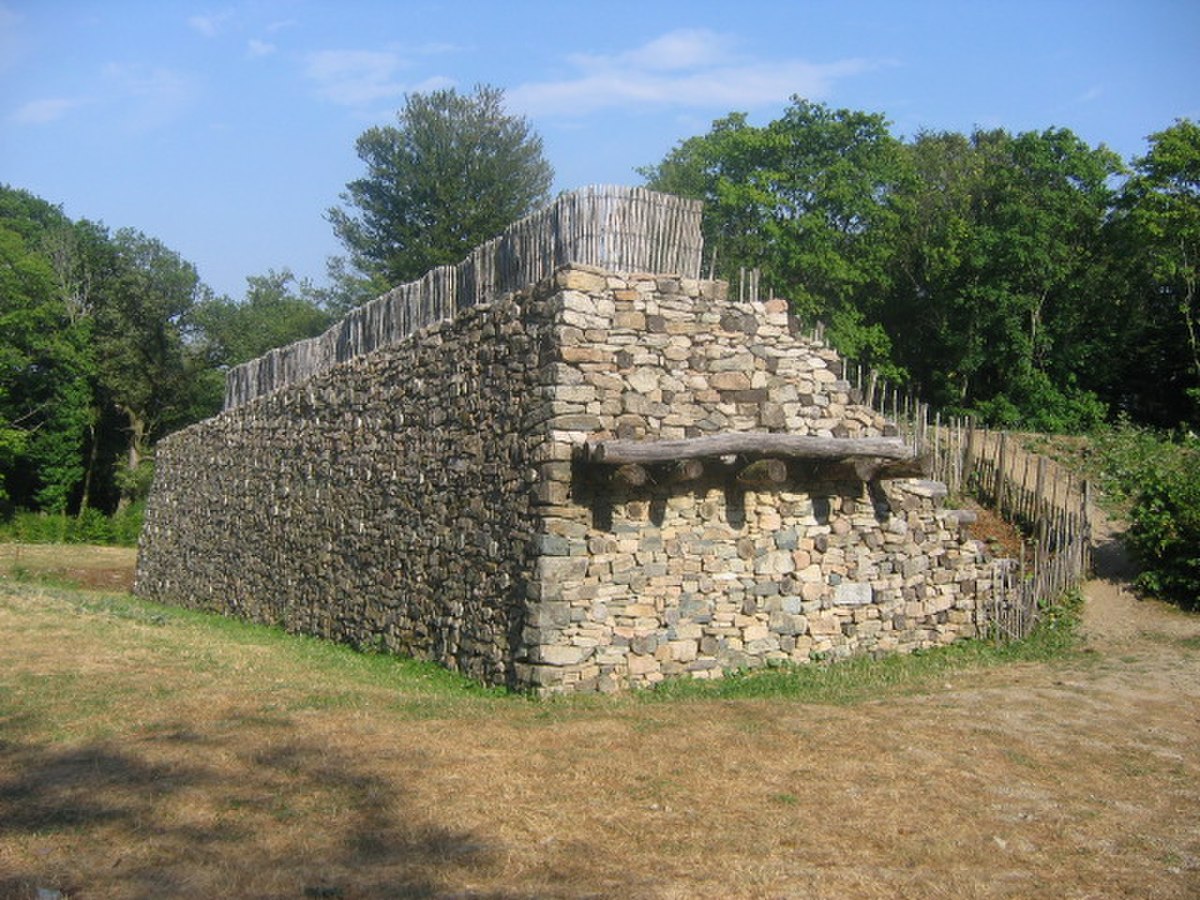 A reconstruction of the walls of Bibracte, a Gaulish oppidum, showcasing the murus gallicus construction technique. The wall is made of stacked stones and timber, with a wooden palisade on top, set in a natural, wooded environment.