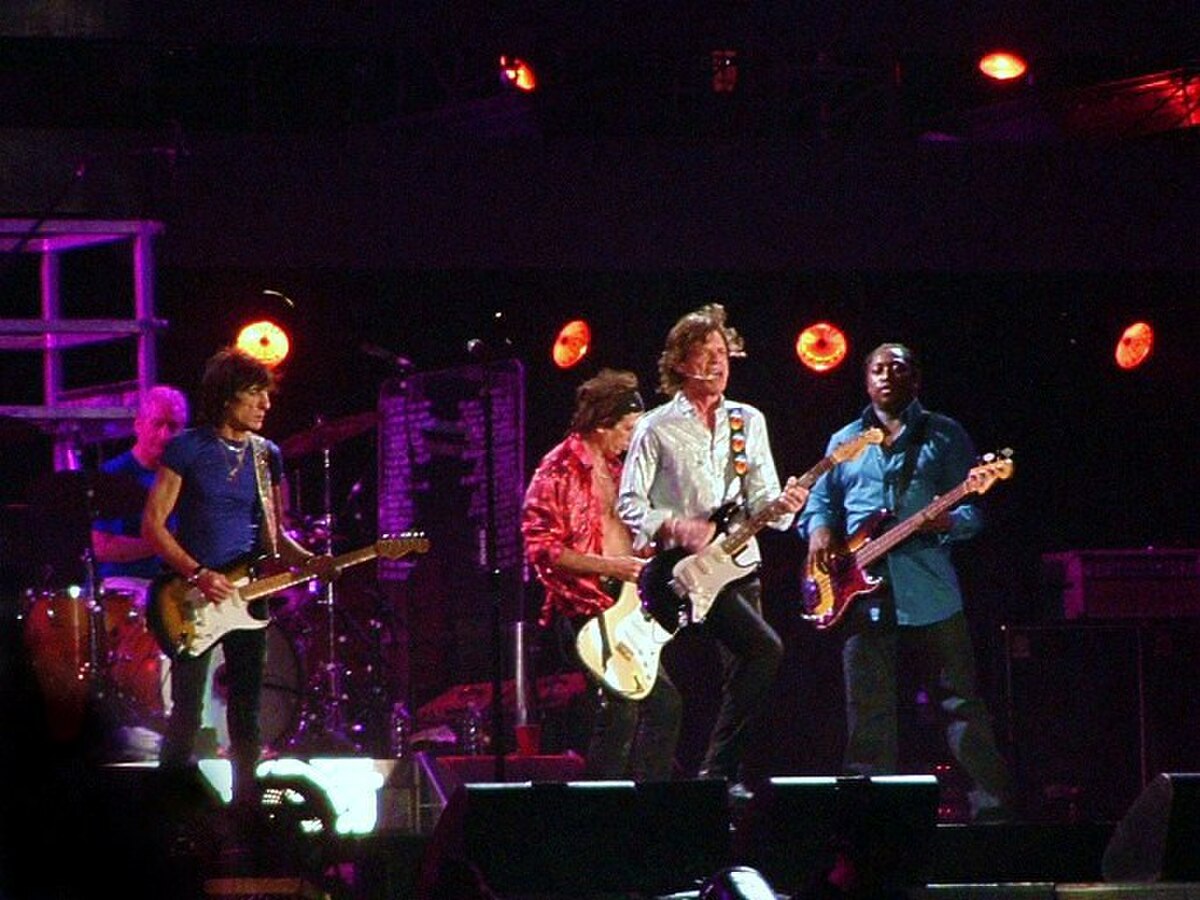 The Rolling Stones perform on stage during a 2006 concert, with several band members playing guitars and bass. Bright stage lights illuminate the performers.