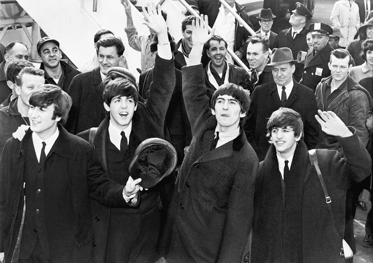 The Beatles arrive at JFK Airport in 1964, with John Lennon, Paul McCartney, George Harrison, and Ringo Starr waving to the crowd. They are surrounded by other people, some of whom are also waving.