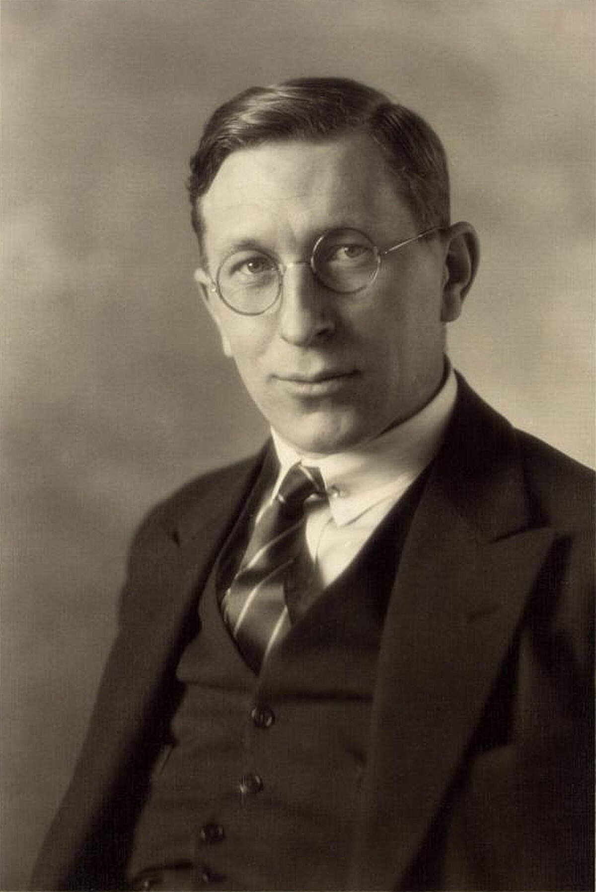 A sepia-toned portrait of Frederick G. Banting from 1923, showing him wearing a suit, tie, and round spectacles. He is looking directly at the viewer with a slight smile.