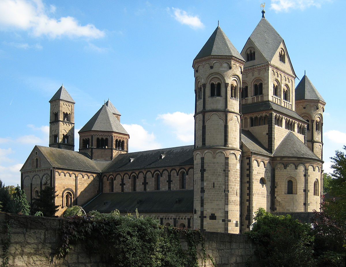 The Maria Laach Abbey, a large Romanesque monastery church with multiple towers and rounded apses, constructed from dark stone. The building is surrounded by trees and set under a partly cloudy sky.