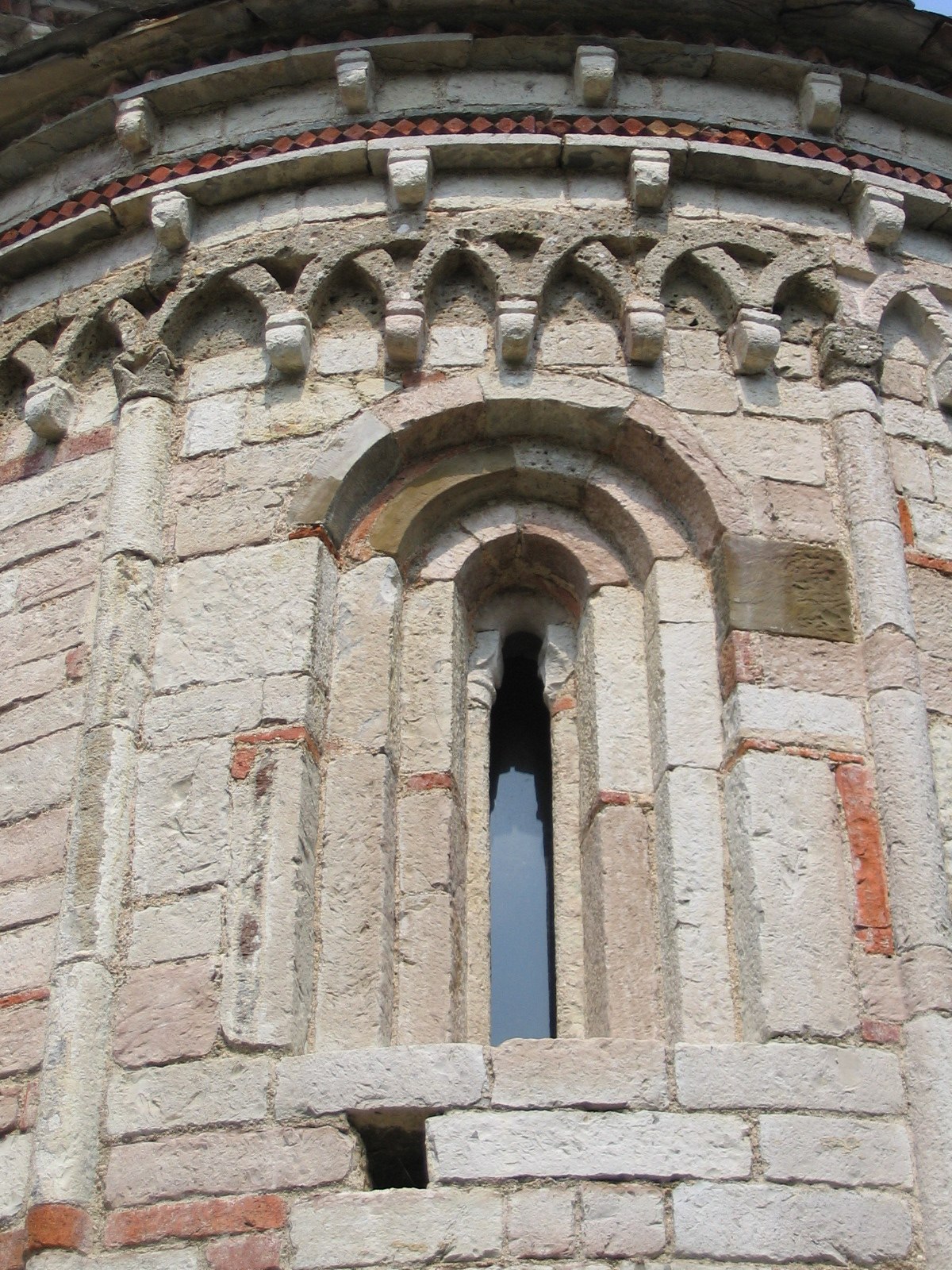 Close-up of a Romanesque arched window set in a stone wall, featuring decorative carvings and a narrow opening. The stonework includes various patterns and textures, indicative of medieval craftsmanship.