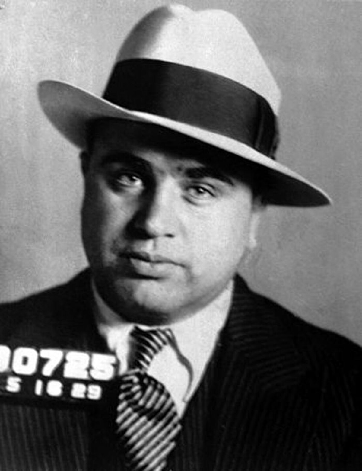 Mugshot of Al Capone, taken by the Bureau of Investigation in 1929, showing him wearing a light-colored fedora and a striped tie. The number "0725" is visible on a card in the lower left.