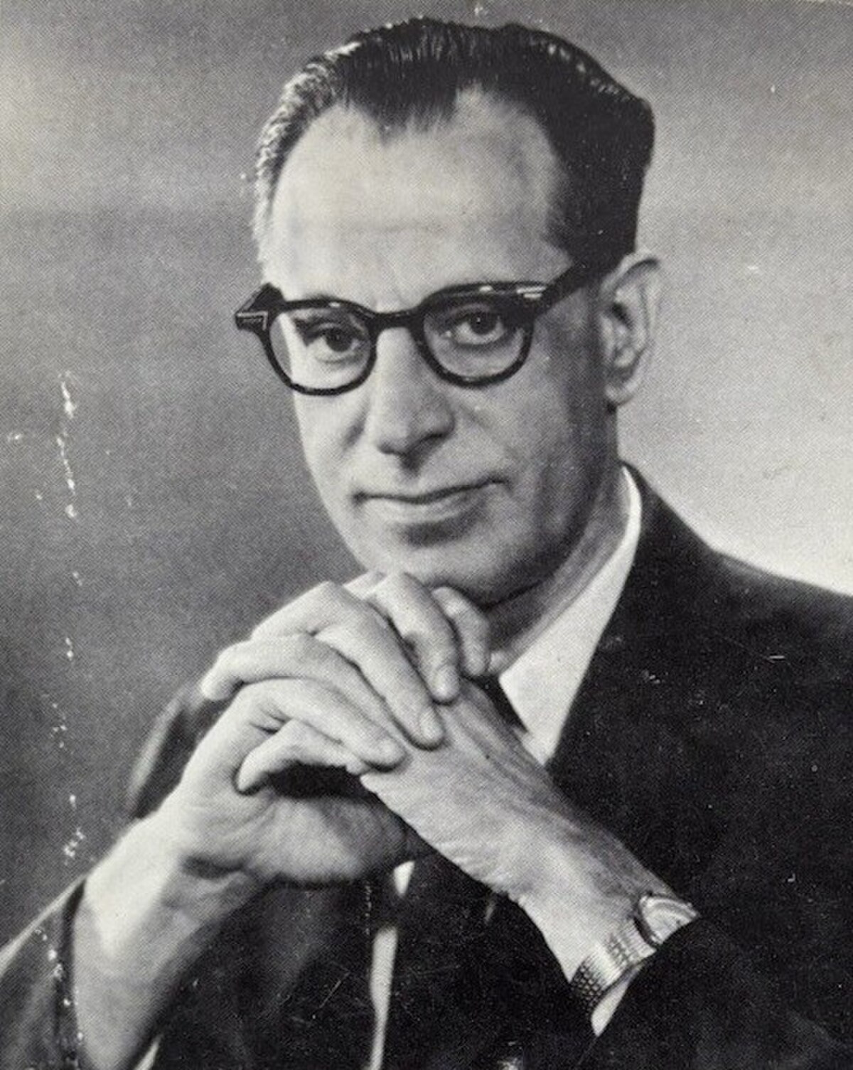 A black and white portrait of Albert Ellis, showing him wearing glasses and a suit, with his hands clasped under his chin. He looks directly at the viewer with a thoughtful expression.