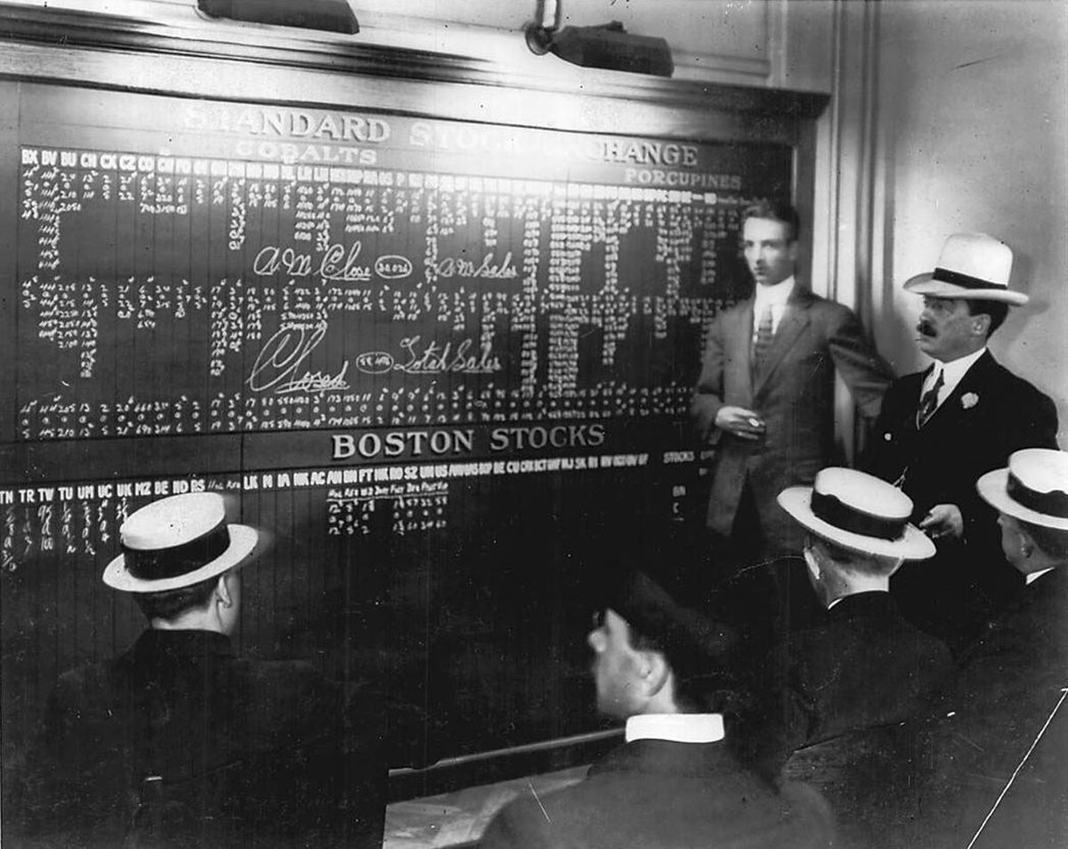 A black and white photograph of the Toronto Stock Exchange in 1910, showing a large board with stock listings and several men in suits and hats. One man points to the board, which lists "STANDARD STOCKS" and "BOSTON STOCKS."