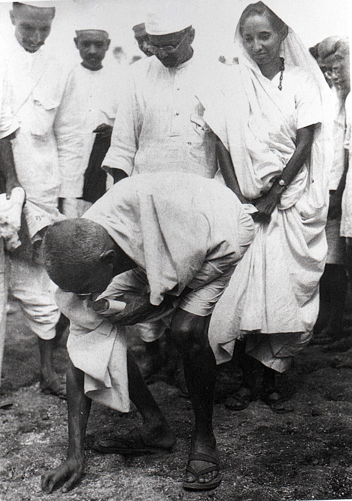 Mahatma Gandhi bends down to collect salt at Dandi Beach on April 6, 1930, during the Salt March. His second son, Manilal Gandhi, and Mithuben Petit stand behind him.