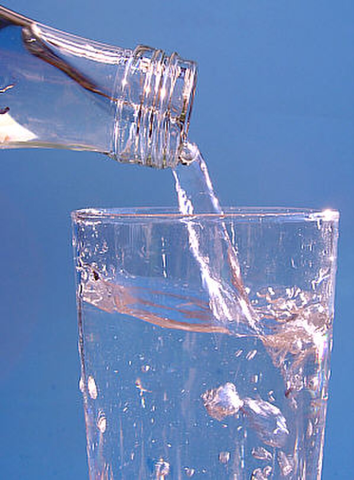 Water is being poured from a clear glass bottle into a clear glass, creating splashes and bubbles. The background is a solid blue color.