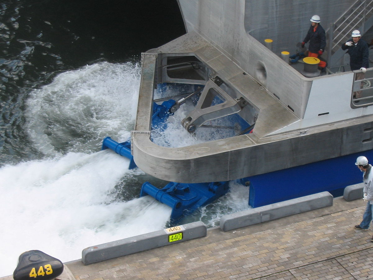 A pump-jet propulsion system is visible at the stern of the NatchanWorld ferry, churning water as it operates. Two workers in hard hats are visible on the deck above the water, and a yellow bollard labeled "443" is on the dock.