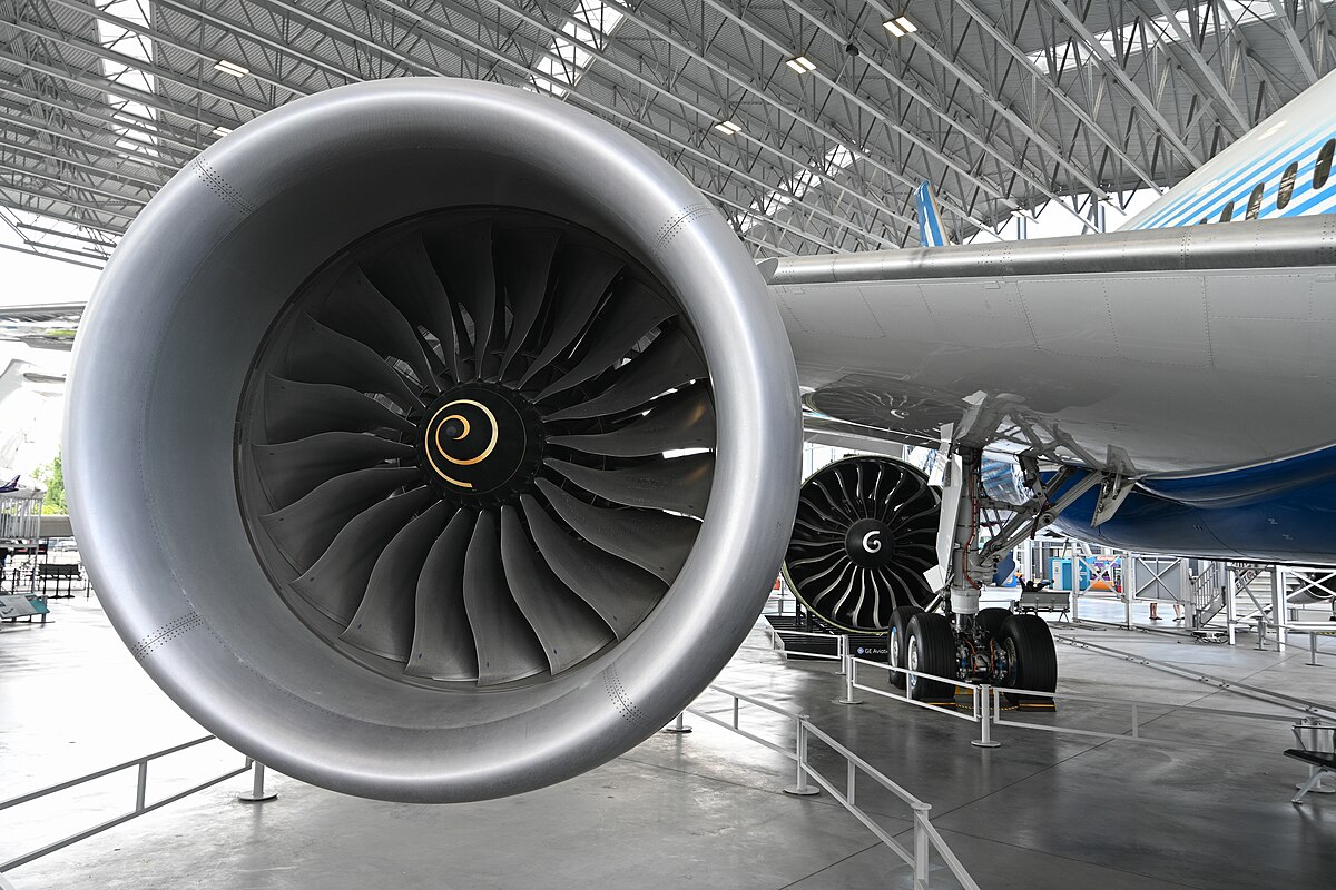 The large Rolls-Royce Trent 1000 jet engine of a Boeing 787 Dreamliner is shown, with its fan blades visible at the front. Another engine and the landing gear are partially visible in the background, inside a hangar.