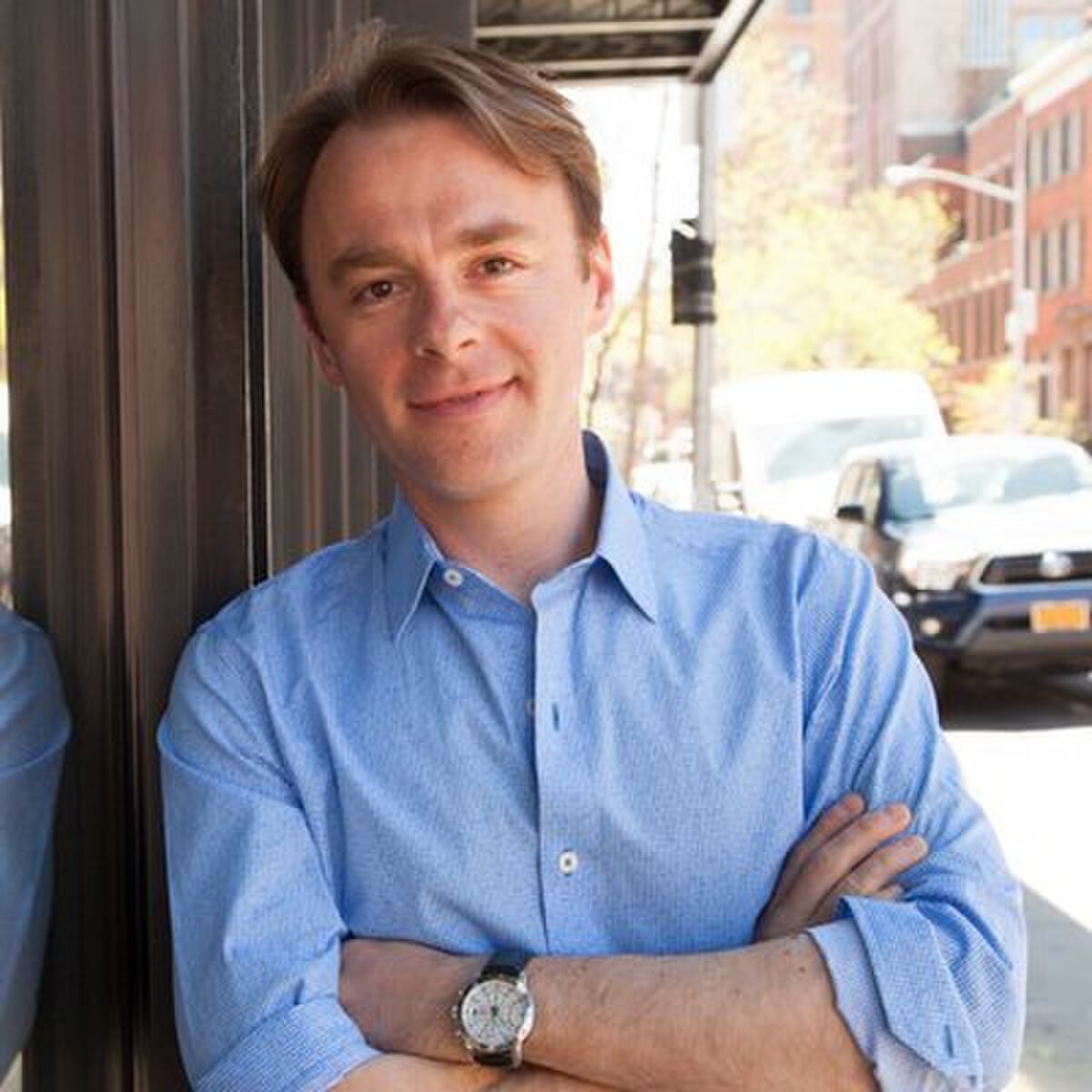 Portrait of Patrick J. McGinnis, who coined the term FOMO, smiling and leaning against a building outdoors. He is wearing a light blue collared shirt and has his arms crossed.