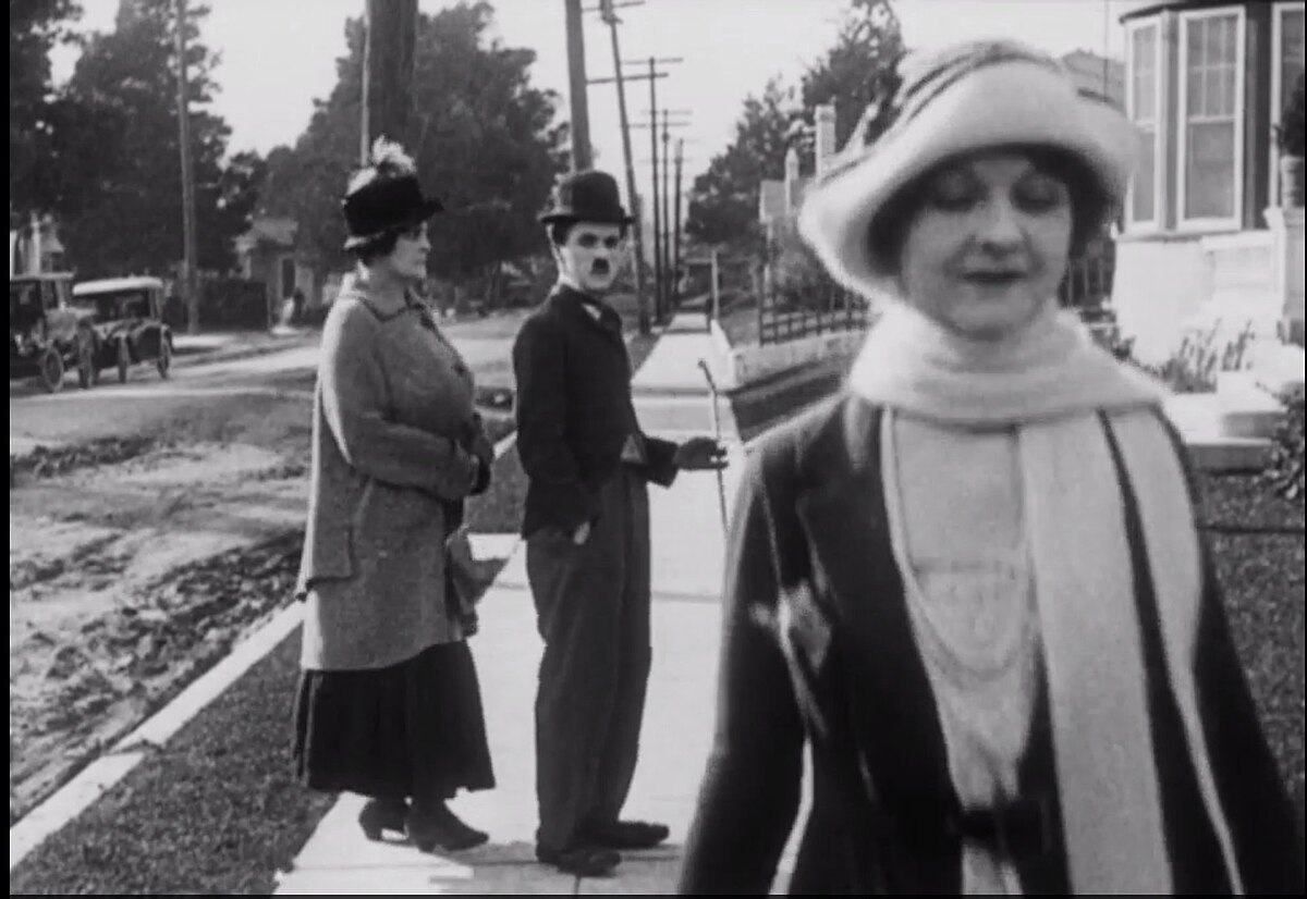A black and white still from Charlie Chaplin's 1922 film "Pay Day," showing Chaplin's character looking back at a woman in the foreground while walking with another woman. The scene captures a moment of distraction, similar to the popular 'distracted boyfriend' meme.