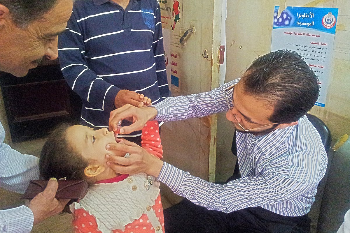 A medical professional administers polio vaccination drops to a young child in Egypt. The child is seated on an adult's lap, and another adult is observing the process.