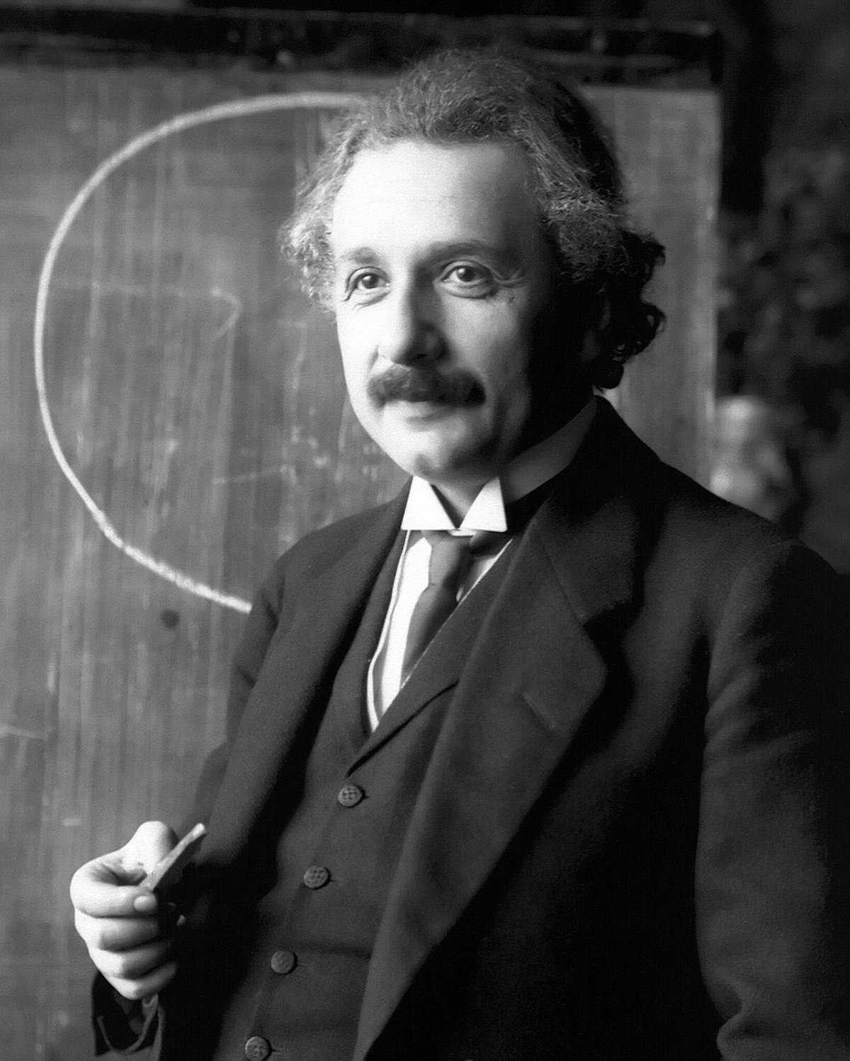 Black and white portrait of Albert Einstein in 1921, holding a piece of chalk in front of a blackboard with a circle drawn on it.
