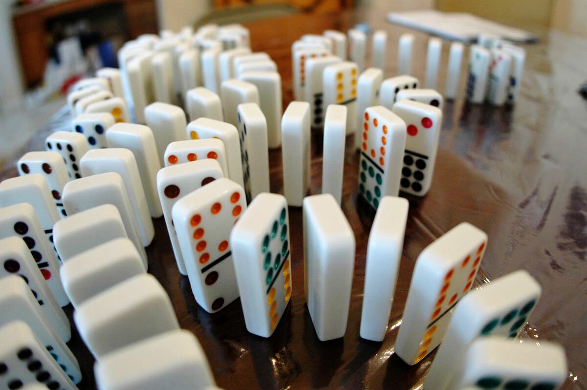 Numerous dominoes, some white with colored dots and others solid white, are arranged in a curved pattern on a table, with some already toppled. The image illustrates the concept of a domino effect.