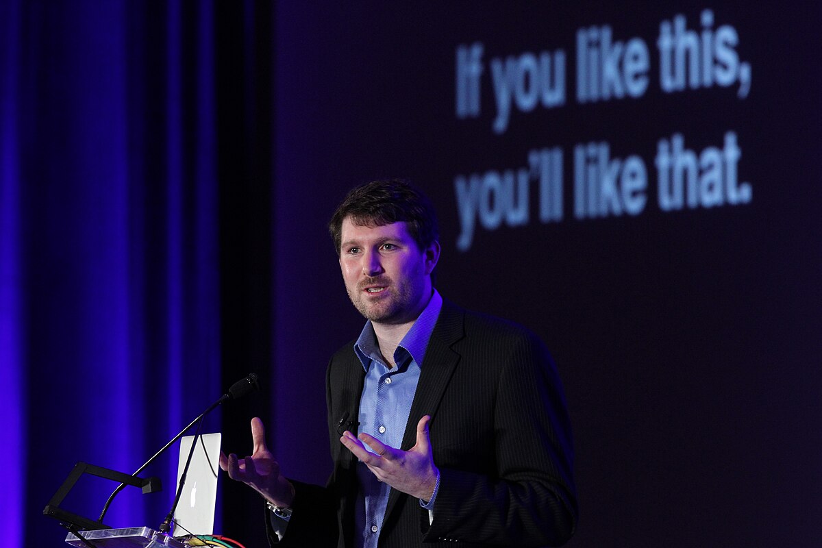 A portrait of Eli Pariser, author of "The Filter Bubble", speaking at a podium with a screen behind him displaying the text "If you like this, you'll like that.". He is wearing a dark jacket over a blue collared shirt and is gesturing with both hands.