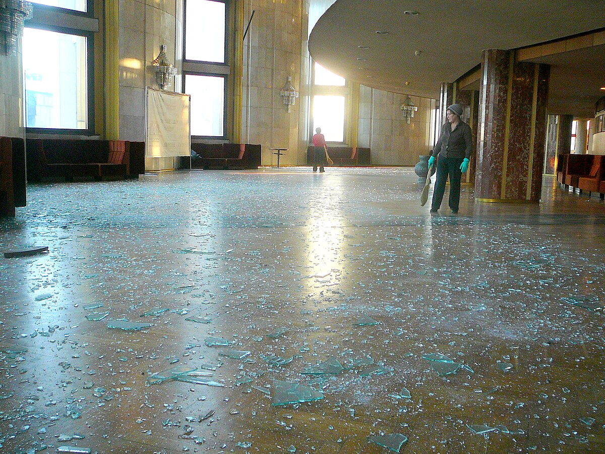 The interior of the Drama Theatre in Chelyabinsk shows extensive damage caused by the shock wave from a meteor event. The floor is covered with shattered glass from broken windows, and two people are visible in the background, assessing the damage.