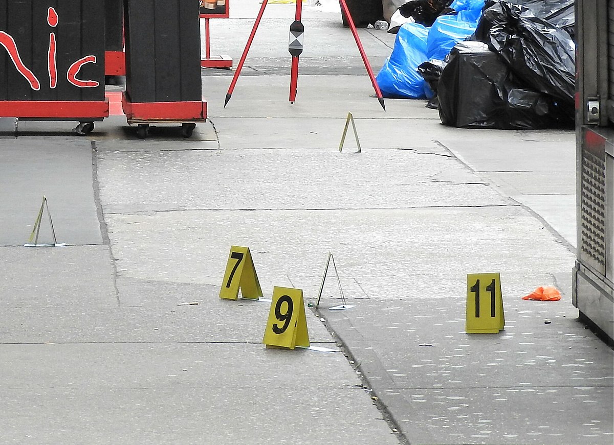 Crime scene with numbered evidence markers on a sidewalk in Manhattan, New York City in December 2017. Markers 7, 9, and 11 are visible, along with a red tripod and black trash bags.