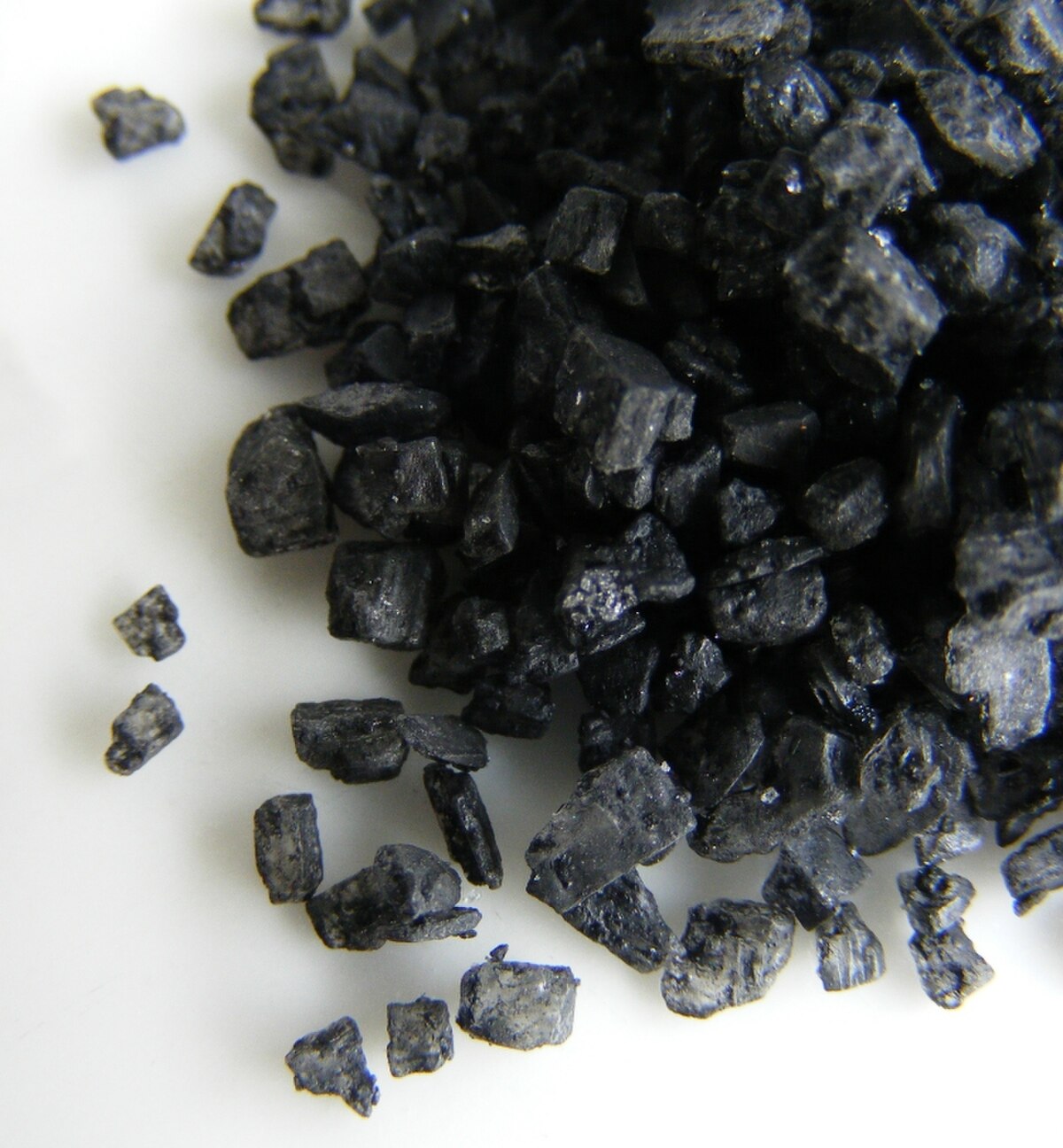 Close-up photograph of black lava salt crystals. The salt crystals are irregularly shaped and dark, with a slightly glossy appearance, scattered on a white surface.