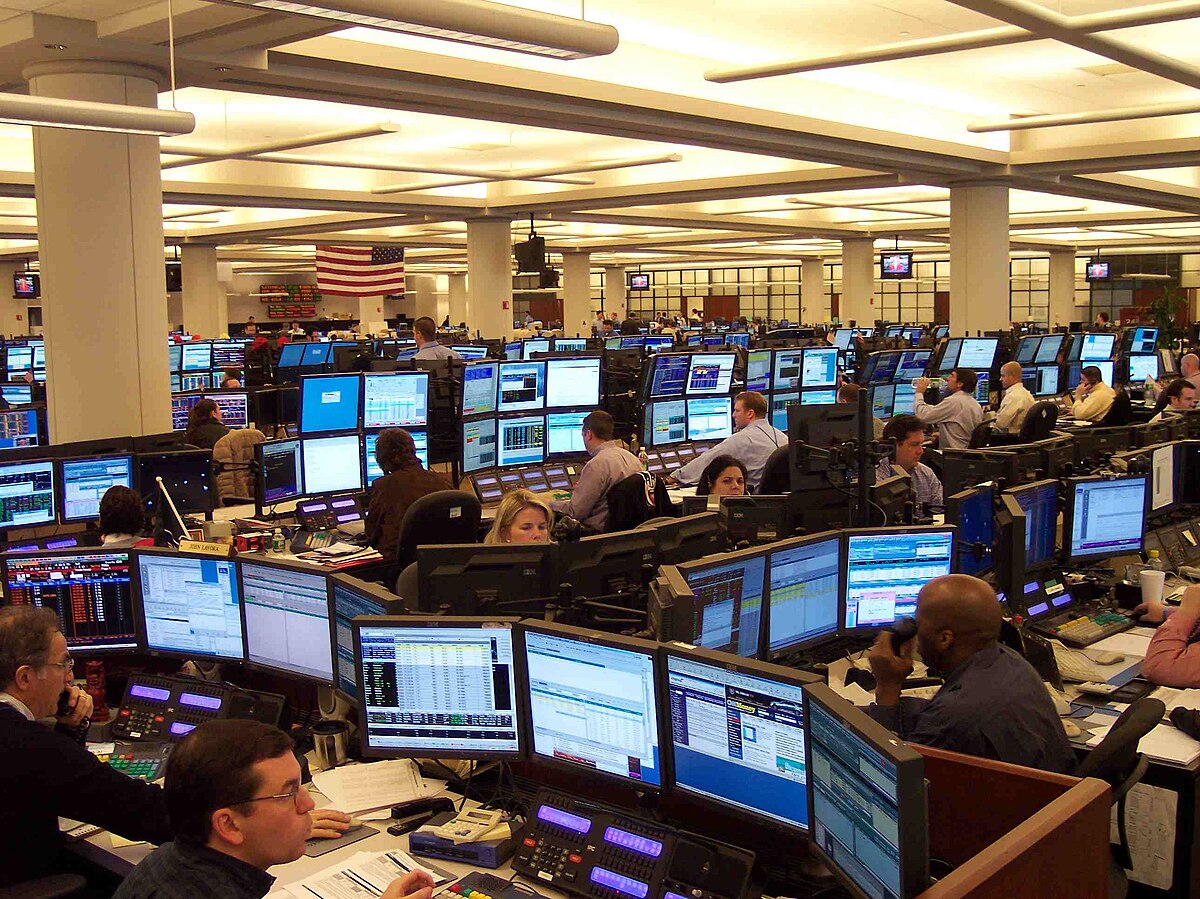 A busy trading floor in Houston in 2009, filled with numerous oil traders seated at desks, each monitoring multiple computer screens. An American flag is visible in the background.