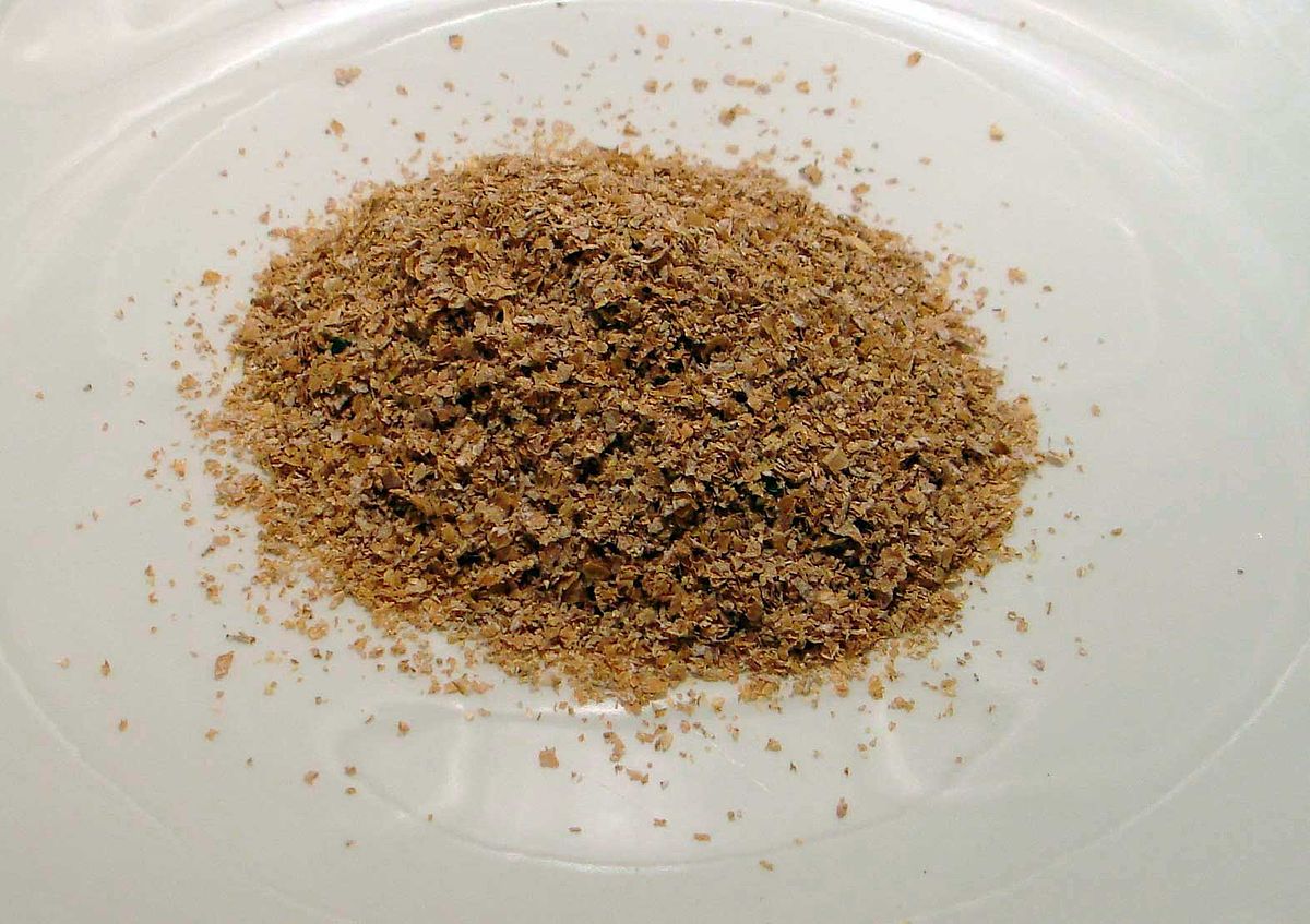 A pile of wheat bran, a food product known for its high content of dietary fiber, is shown on a white plate. The bran consists of small, brown, flaky particles.