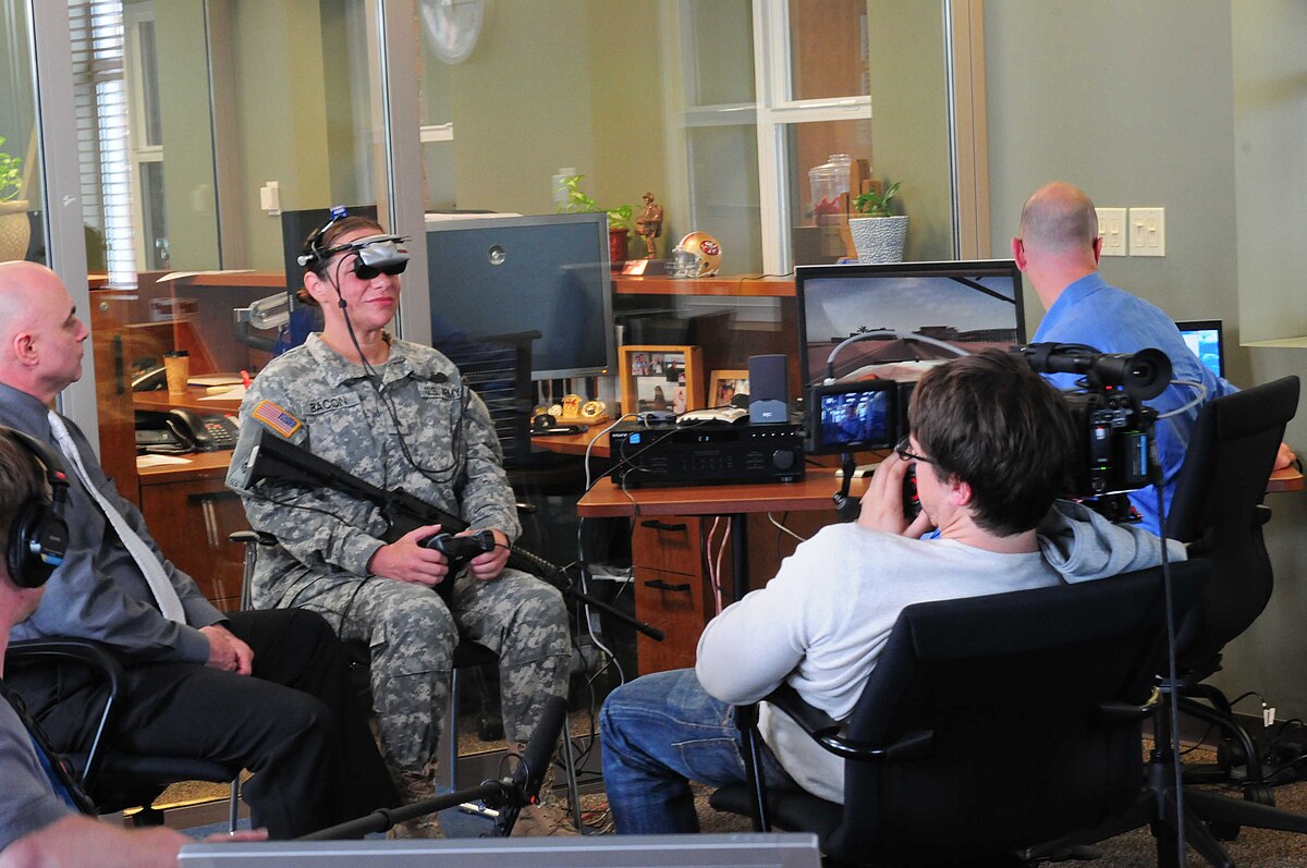 A soldier in uniform wearing a virtual reality headset and holding a rifle controller participates in exposure therapy for PTSD at Joint Base Lewis-McChord. A documentary crew films the session while monitors display the virtual environment.