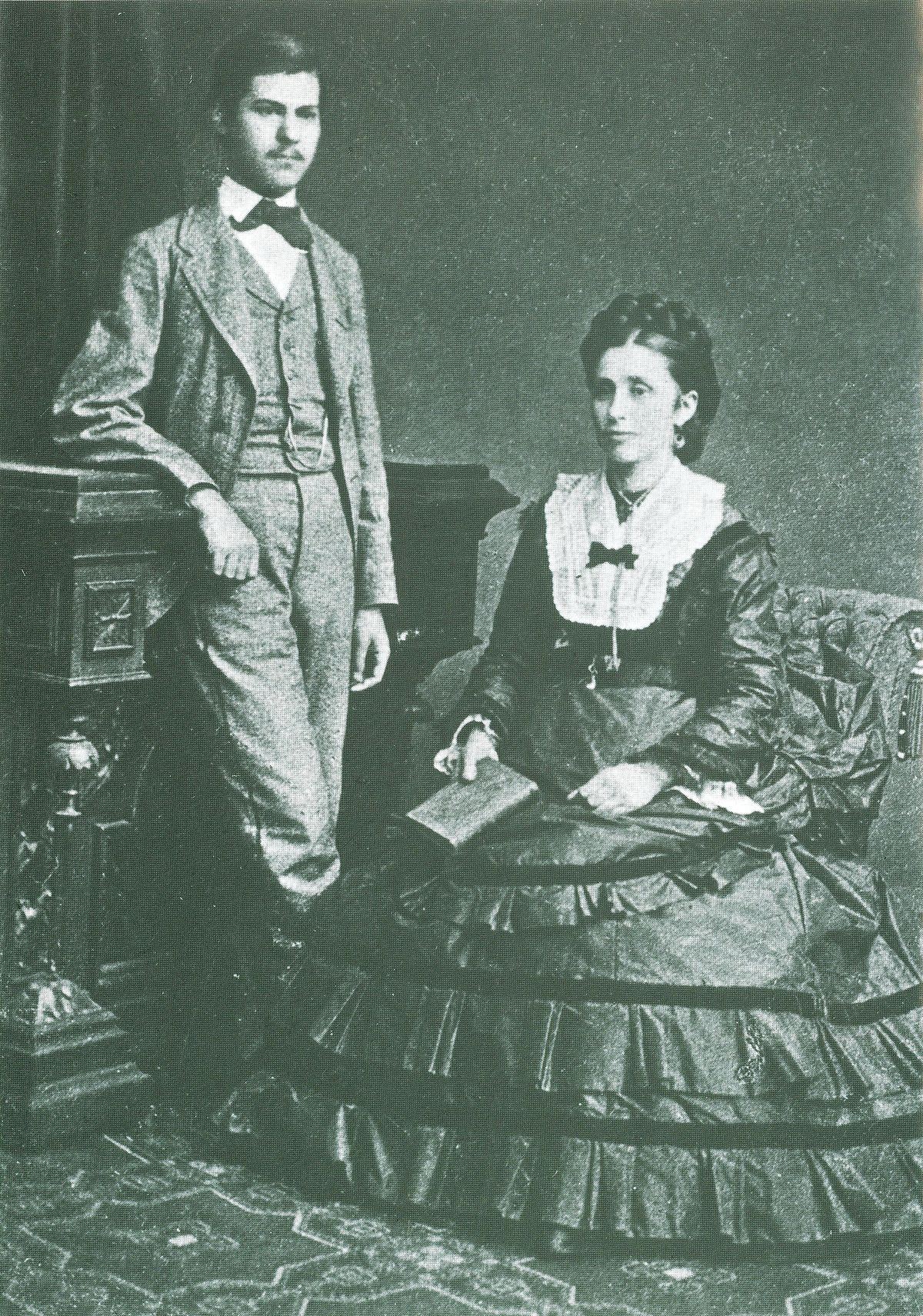 A black and white photograph of a 16-year-old Sigmund Freud with his mother, Amalia Freud, in 1872. Sigmund Freud stands formally dressed next to his seated mother, who holds a book.