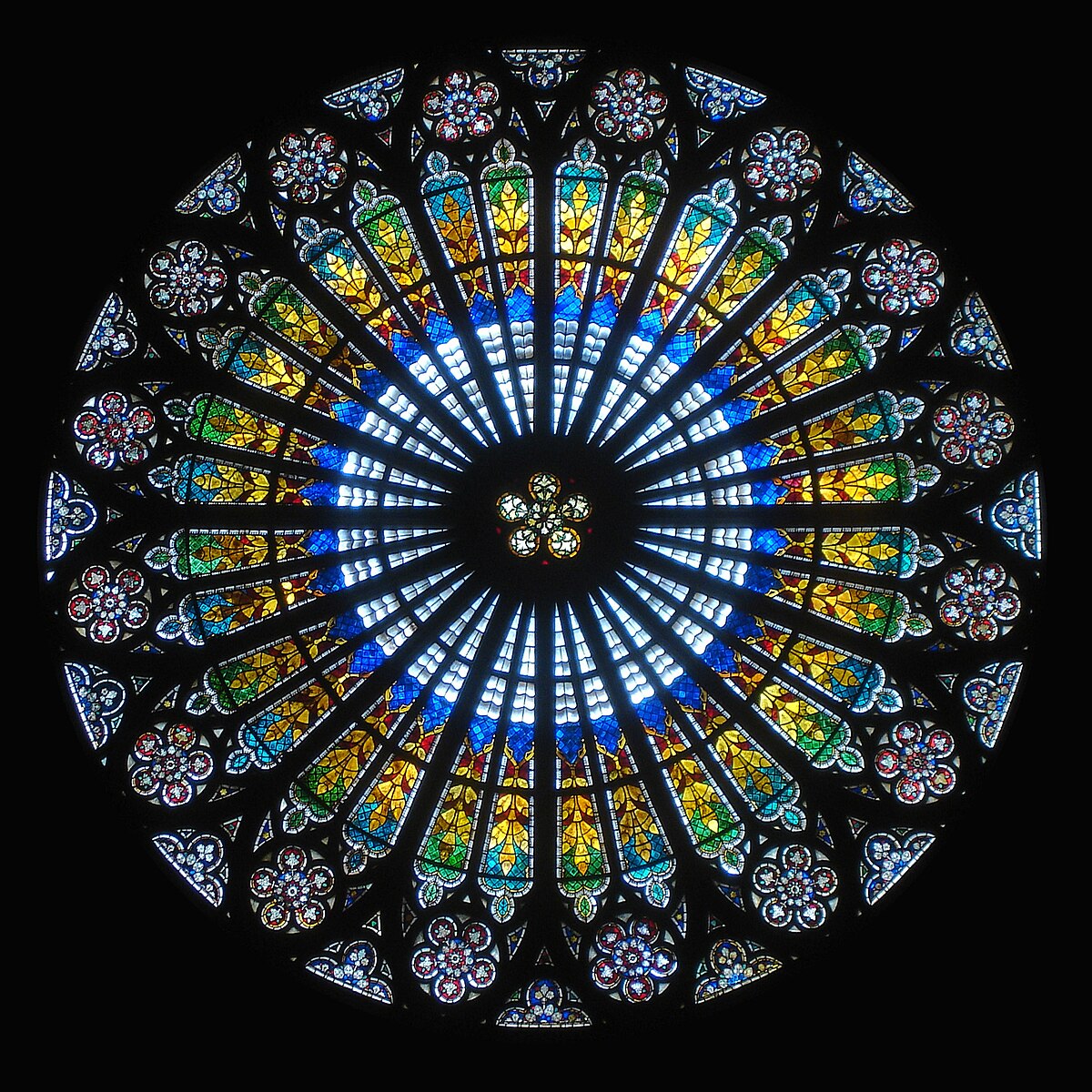 The interior view of the rose window at Strasbourg Cathedral displays vibrant stained glass in a radial pattern. The window features a central star-like motif surrounded by concentric rings of blue, green, yellow, and red glass, forming intricate floral and geometric designs.