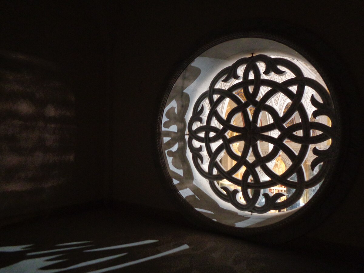 A rose window with an intricate geometric pattern is seen in shadow, casting light patterns on the surrounding wall inside the Church of Saint Sava in Belgrade. The circular window features interwoven loops and curves.