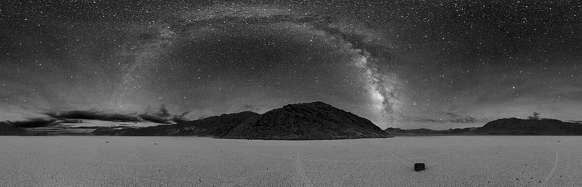 A black and white 360° panoramic photograph of the Milky Way arching across the night sky over a desert landscape in Death Valley. The foreground shows a vast, flat expanse, with mountains and hills silhouetted against the starry sky.